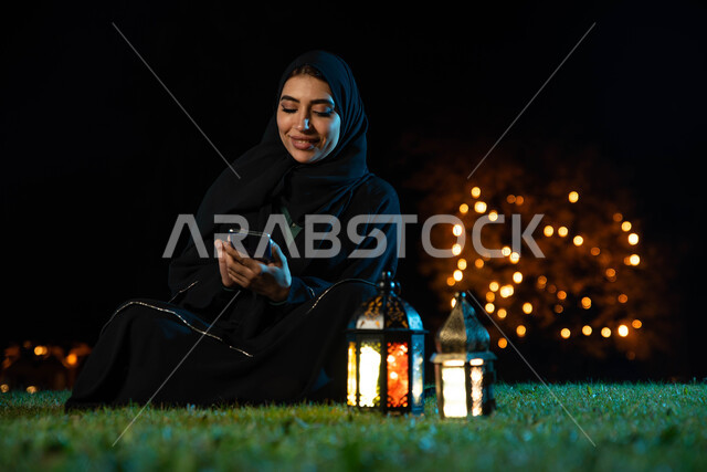 التواصل الاجتماعي وتصفح المواقع الالكترونية، شابة عربية خليجية سعودية ترتدي  العباءة الخليجية تستخدم الهاتف المحمول تجلس في الهواء الطلق في الخارج ، فانوس رمضان، شهر رمضان المبارك وإضاءة وزينة شهر رمضان، أجواء رمضانية