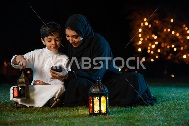 استخدام الهاتف المحمول وتصفح المواقع الالكترونية، أمرأة عربية خليجية سعودية ترتدي العباءة الخليجية تقضي اوقات ممتعة مع ابنها في الهواء الطلق في الخارج ، فانوس رمضان، إضاءة وزينة شهر رمضان