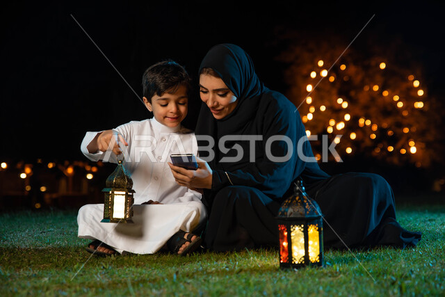 استخدام الهاتف المحمول وتصفح المواقع الالكترونية، أم عربية خليجية سعودية ترتدي العباءة الخليجية تقضي اوقات ممتعة مع ابنها في الهواء الطلق في الخارج ، فانوس رمضان، إضاءة وزينة شهر رمضان