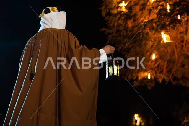 أجواء رمضانية، مناسبات دينية ، اسلاميات وعبادات ، صورة من الخلف لرجل عربي سعودي خليجي يرتدي الزي التراثي الشعبي التقليدي يمسك بيده فانوس رمضان ، النظر الى شيء ما، يوم التاسيس السعودي ، اليوم الوطني السعودي، تراثيات وشعبيات ، إضاءة وزينة شهر رمضان