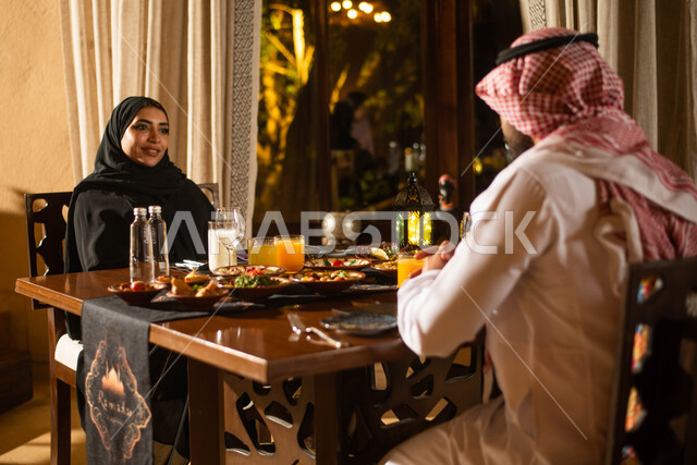 A Saudi Arabian Gulf couple at the breakfast table in the month of Ramadan, decorations for the month of Ramadan and a trip full of varieties of delicious food and eating breakfast, happy facial gestures and Ramadan atmosphere, Ramadan lantern, the holy m