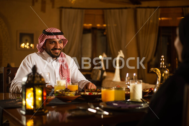 سفرة مليئة باصناف من المأكولات اللذيذة وتناول وجبة الافطار ، زوجان عربيان خليجيان سعوديان على مائدة الافطار بشهر رمضان ، ايماءات وجه تدل على السعادة والسرور ، اجواء رمضانية، فانوس رمضان، شهر رمضان الكريم
