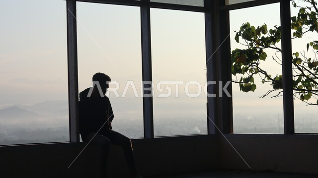 صورة ظلية لرجل يجلس امام النافذة  داخل المبنى, النظر وتأمل الطبيعه من النافذه,  مناظر طبيعيه من الأبراج والمباني المرتفعه , التفكير والإنعزال عند النافذه, سيلوليت