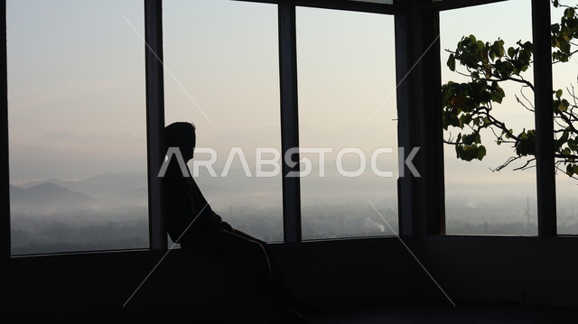 صورة ظلية لرجل يجلس امام النافذة  داخل المبنى, النظر وتأمل الطبيعه من النافذه,  مناظر طبيعيه من الأبراج والمباني المرتفعه , التفكير والإنعزال عند النافذه, سيلوليت