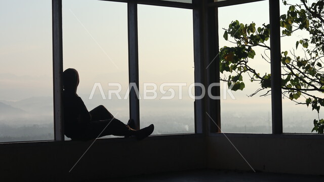 صورة ظلية لرجل يجلس امام النافذة  داخل المبنى, النظر وتأمل الطبيعه من النافذه,  مناظر طبيعيه من الأبراج والمباني المرتفعه , التفكير والإنعزال عند النافذه, سيلوليت