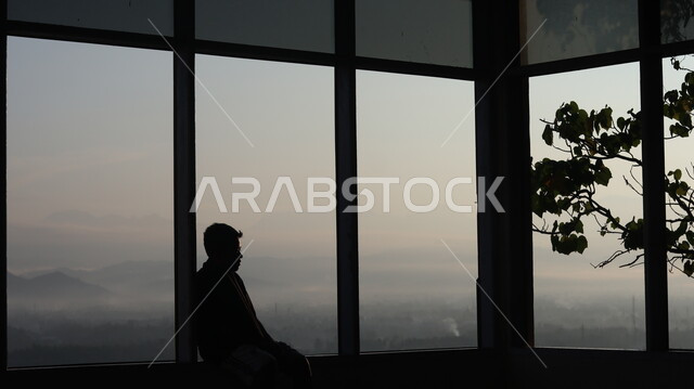 صورة ظلية لرجل يجلس امام النافذة  داخل المبنى, النظر وتأمل الطبيعه من النافذه,  مناظر طبيعيه من الأبراج والمباني المرتفعه , التفكير والإنعزال عند النافذه, سيلوليت