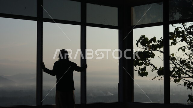 صورة ظلية لرجل يقف امام النافذة  داخل المبنى, النظر وتأمل الطبيعه من النافذه,  مناظر طبيعيه من الأبراج والمباني المرتفعه , التفكير والإنعزال عند النافذه, سيلوليت