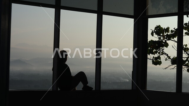 صورة ظلية لرجل يجلس امام النافذة  داخل المبنى, النظر وتأمل الطبيعه من النافذه,  مناظر طبيعيه من الأبراج والمباني المرتفعه , التفكير والإنعزال عند النافذه, سيلوليت