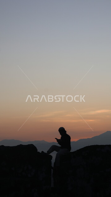 استخدام أجهزة تقنية حديثة, الاسترخاء والتأمل , صورة ظلية  لشاب يجلس على الصخور في المرتفعات الجبلية ويستخدم الهاتف المحمول , السماء والمناظر الطبيعية , منظر غروب الشمس ، خلفية الطبيعة , سيلوليت