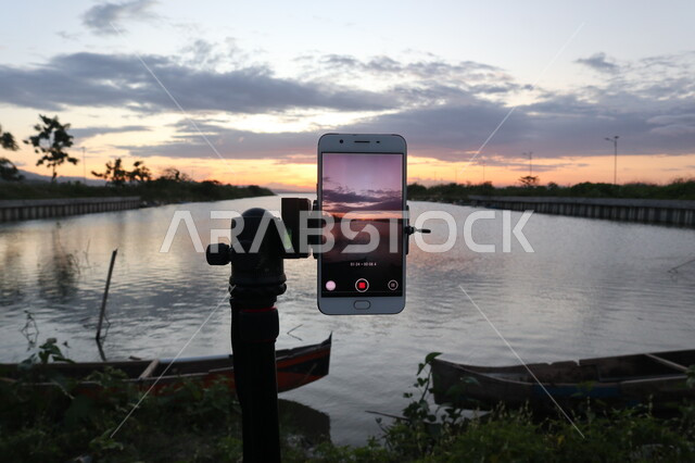 هاتف محمول على حامل تثبيت الهواتق والكاميرات، التقاط صورة تذكارية من خلال الكاميرا الخلفية للهاتف المحمول، الاشجار والنباتات الخضراء المحيطة بالبحيرة المائية، مناظر طبيعية، خلفية الطبيعة