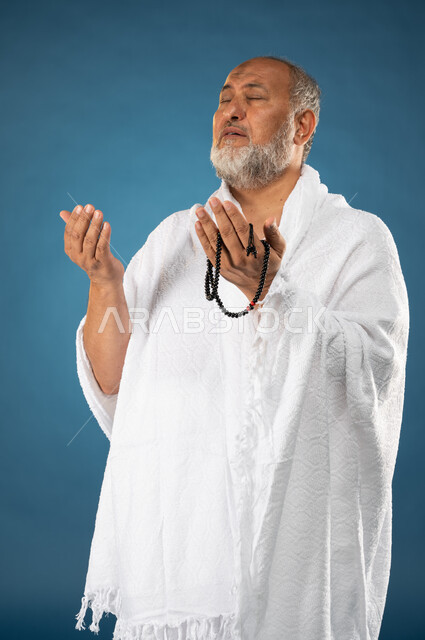 الدعاء والعباده والتقرب الى الله، بورتريه لرجل مسن مسلم عربي سعودي خليجي يرتدي ملابس الاحرام ويمسك بيده مسبحة ويرفع يديه لاعلى، آداء مناسك الحج والعمرة، الركن الخامس من أركان الاسلام، موسم الحج 2023، خلفية زرقاء