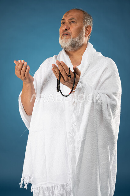 الدعاء والعباده والتقرب الى الله، بورتريه لرجل مسن مسلم عربي سعودي خليجي يرتدي ملابس الاحرام ويمسك بيده مسبحة ويرفع يديه لاعلى، آداء مناسك الحج والعمرة، الركن الخامس من أركان الاسلام، موسم الحج 2023، خلفية زرقاء