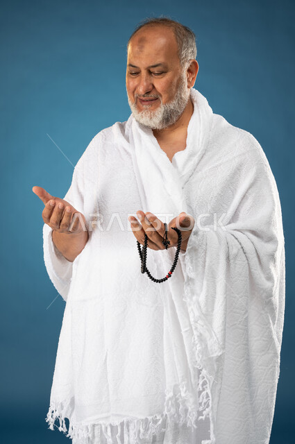 الدعاء والعباده والتقرب الى الله، بورتريه لرجل مسن مسلم عربي سعودي خليجي يرتدي ملابس الاحرام ويمسك بيده مسبحة ويرفع يديه لاعلى، آداء مناسك الحج والعمرة، الركن الخامس من أركان الاسلام، موسم الحج 2023، خلفية زرقاء