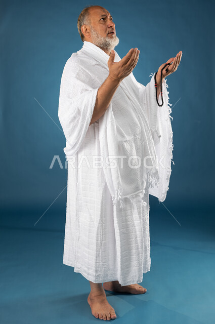 Performing the rituals of Hajj and Umrah, wearing Ihram clothes, a portrait of an elderly Muslim, Arab, Saudi, Gulf man holding a rosary, raising his hands up, supplication, worship, and drawing closer to God, the fifth pillar of Islam, Hajj season 2023, 