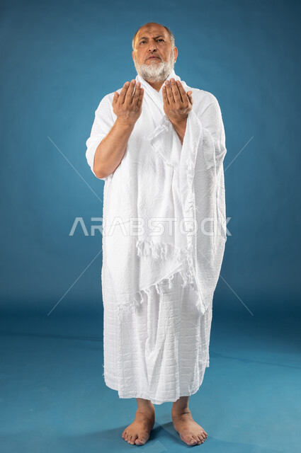 Wearing ihram clothes, a portrait of an elderly, Arab, Saudi, Gulf, Muslim man raising his hands up, supplication, worship, and drawing closer to God, performing the rituals of Hajj and Umrah, the fifth pillar of Islam, Hajj season 2023, full-length body 