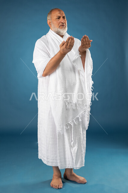ارتداء ملابس الاحرام، بورتريه لرجل مسن مسلم عربي سعودي خليجي يرفع يديه لاعلى، الدعاء والعباده والتقرب الى الله، آداء مناسك الحج والعمرة، الركن الخامس من أركان الاسلام، موسم الحج 2023، صورة الجسم بالطول الكامل، خلفية زرقاء