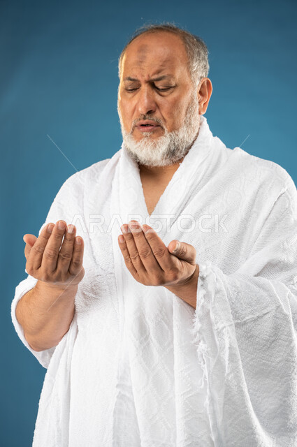ارتداء ملابس الاحرام، بورتريه لرجل مسن مسلم عربي سعودي خليجي يرفع يديه لاعلى، الدعاء والعباده والتقرب الى الله، آداء مناسك الحج والعمرة، الركن الخامس من أركان الاسلام، موسم الحج 2023، خلفية زرقاء