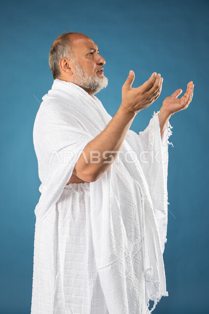 ارتداء ملابس الاحرام، بورتريه لرجل مسن مسلم عربي سعودي خليجي يرفع يديه لاعلى، الدعاء والعباده والتقرب الى الله، آداء مناسك الحج والعمرة، الركن الخامس من أركان الاسلام، موسم الحج 2023، خلفية زرقاء