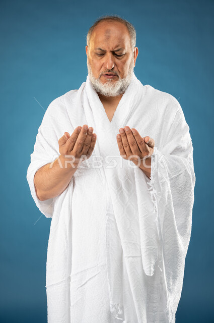 ارتداء ملابس الاحرام، بورتريه لرجل مسن مسلم عربي سعودي خليجي يرفع يديه لاعلى، الدعاء والعباده والتقرب الى الله، آداء مناسك الحج والعمرة، الركن الخامس من أركان الاسلام، موسم الحج 2023، خلفية زرقاء