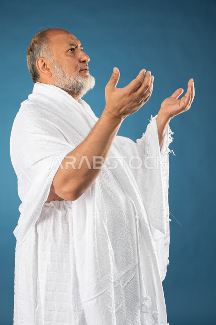 الدعاء والعباده والتقرب الى الله، بورتريه لرجل مسن مسلم عربي سعودي خليجي يرتدي ملابس الاحرام يرفع يديه لاعلى، آداء مناسك الحج والعمرة، الركن الخامس من أركان الاسلام، موسم الحج 2023، خلفية زرقاء