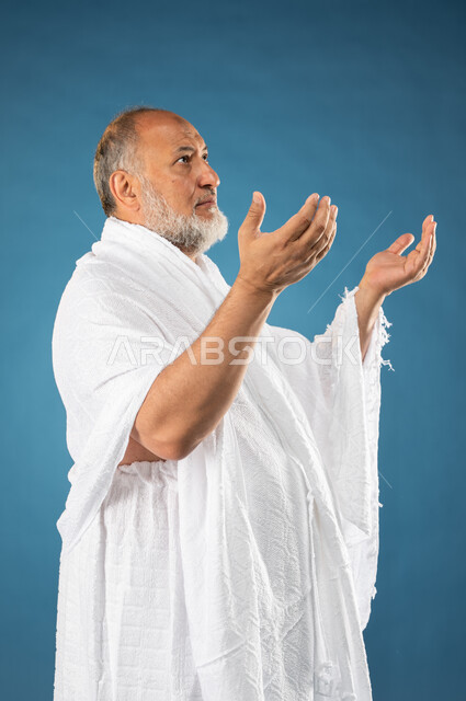 الدعاء والعباده والتقرب الى الله، بورتريه لرجل مسن مسلم عربي سعودي خليجي يرتدي ملابس الاحرام يرفع يديه لاعلى، آداء مناسك الحج والعمرة، الركن الخامس من أركان الاسلام، موسم الحج 2023، خلفية زرقاء
