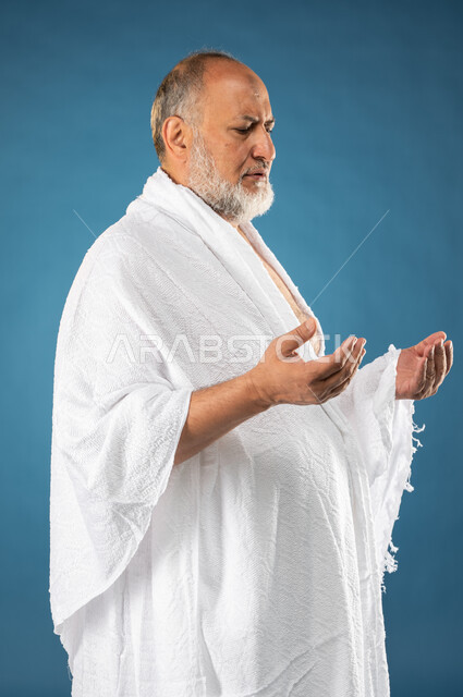الدعاء والعباده والتقرب الى الله، بورتريه لرجل مسن مسلم عربي سعودي خليجي يرتدي ملابس الاحرام يرفع يديه لاعلى، آداء مناسك الحج والعمرة، الركن الخامس من أركان الاسلام، موسم الحج 2023، خلفية زرقاء