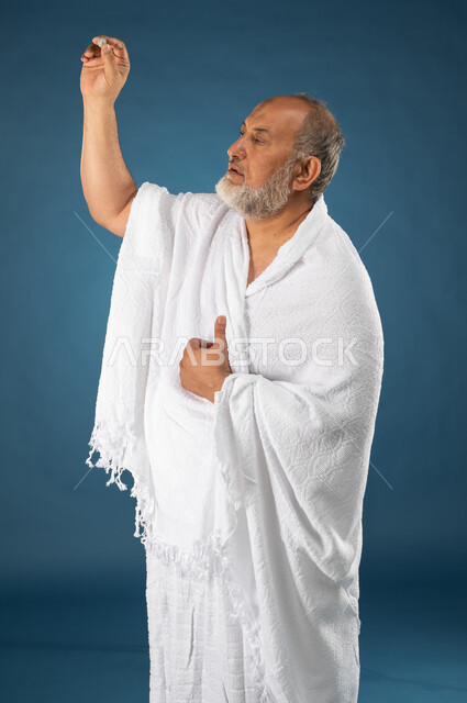 رمي جمرة العقبة في يوم النحر، بورتريه لرجل مسن مسلم عربي سعودي خليجي يرتدي ملابس الاحرام ويمسك بيده حصوى، رمي الحصى ورجم ابليس، العباده والتقرب الى الله، آداء مناسك الحج والعمرة، الركن الخامس من أركان الاسلام، موسم الحج 2023، خلفية زرقاء