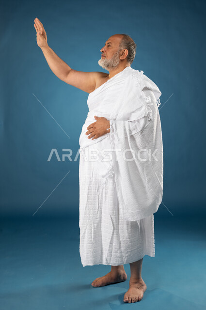 Performing the rituals of Hajj and Umrah, a portrait of an elderly Muslim, Arab, Saudi, and Gulf man wearing Ihram clothes and raising his hand to say hello and peace, worship and drawing closer to God, the fifth pillar of Islam, Hajj season 2023, blue ba