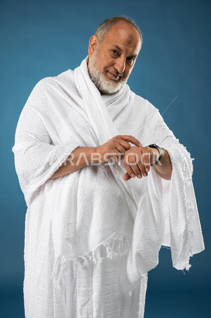 استخدام ساعة يد الكترونية، بورتريه لرجل مسن مسلم  سعودي خليجي عربي يرتدي ملابس الاحرام، معرفة وتحديد الوقت، مفهوم ادارة الوقت، موسم الحج 2023، آداء مناسك الحج والعمرة، الركن الخامس من أركان الاسلام، خلفية زرقاء