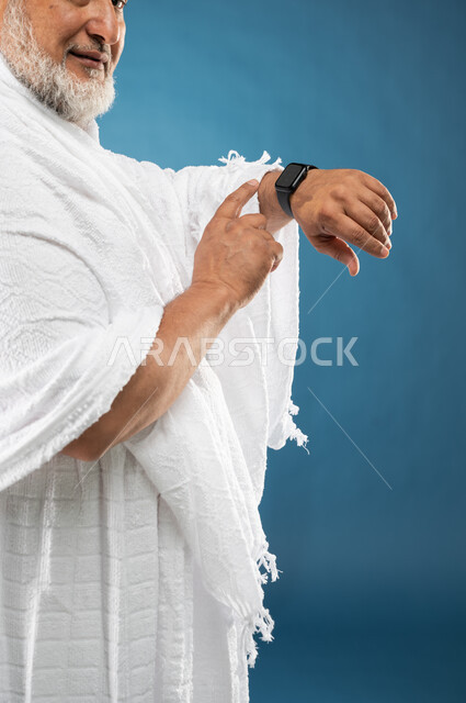 معرفة وتحديد الوقت، مفهوم ادارة الوقت، بورتريه مقرب لرجل مسن مسلم عربي سعودي خليجي يرتدي ملابس الاحرام ويستخدم ساعة يد الكترونية، موسم الحج 2023، آداء مناسك الحج والعمرة، الركن الخامس من أركان الاسلام، خلفية زرقاء