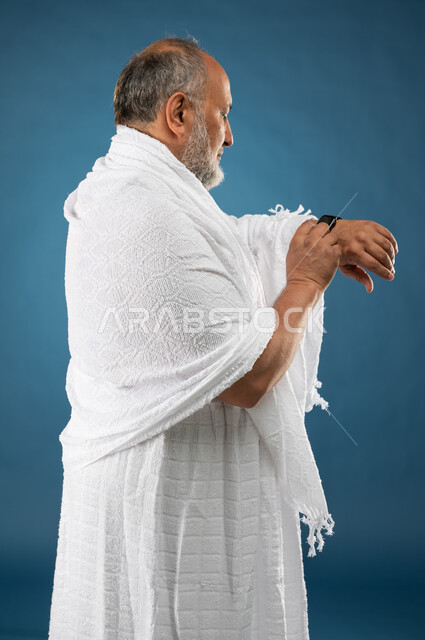 مفهوم ادارة الوقت، بورتريه لرجل مسن مسلم عربي سعودي خليجي يرتدي ملابس الاحرام ويستخدم ساعة يد الكترونية، معرفة وتحديد الوقت، موسم الحج 2023، آداء مناسك الحج والعمرة، الركن الخامس من أركان الاسلام، خلفية زرقاء