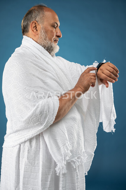مفهوم ادارة الوقت، بورتريه لرجل مسن مسلم عربي سعودي خليجي يرتدي ملابس الاحرام ويستخدم ساعة يد الكترونية، معرفة وتحديد الوقت، موسم الحج 2023، آداء مناسك الحج والعمرة، الركن الخامس من أركان الاسلام، خلفية زرقاء