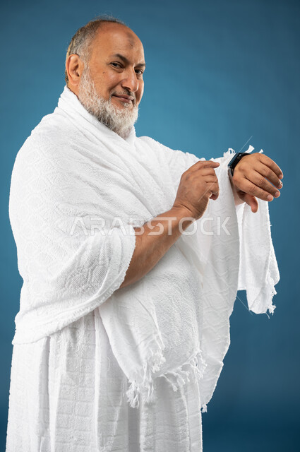 مفهوم ادارة الوقت، بورتريه لرجل مسن مسلم عربي سعودي خليجي يرتدي ملابس الاحرام ويستخدم ساعة يد الكترونية، معرفة وتحديد الوقت، موسم الحج 2023، آداء مناسك الحج والعمرة، الركن الخامس من أركان الاسلام، خلفية زرقاء