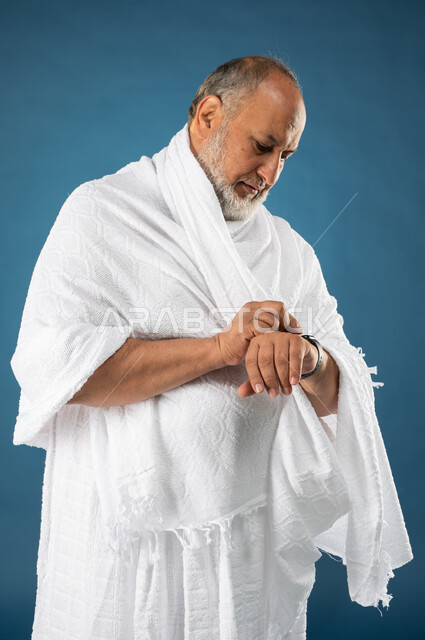 مفهوم ادارة الوقت، بورتريه لرجل مسن مسلم عربي سعودي خليجي يرتدي ملابس الاحرام ويستخدم ساعة يد الكترونية، معرفة وتحديد الوقت، موسم الحج 2023، آداء مناسك الحج والعمرة، الركن الخامس من أركان الاسلام، خلفية زرقاء