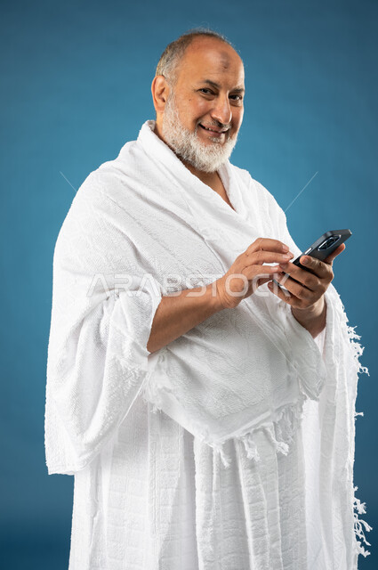 استخدام خدمة تطبيق الارشادات لاداء مناسك الحج والعمرة، بورتريه لرجل مسن مسلم خليجي عربي سعودي يرتدي ملابس الاحرام ويستخدم الهاتف المحمول، تطبيق مرشد الحج الالكتروني، الركن الخامس من أركان الاسلام، موسم الحج 2023، خلفية زرقاء