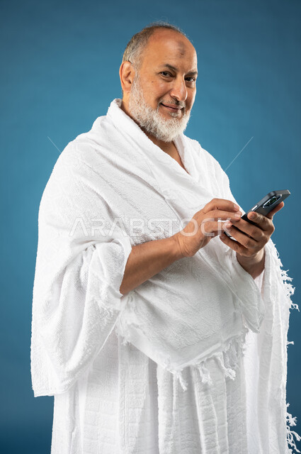 استخدام خدمة تطبيق مرشد الحج الالكتروني، بورتريه لرجل مسن مسلم خليجي عربي سعودي يرتدي ملابس الاحرام ويستخدم الهاتف المحمول، تطبيق الارشادات لاداء مناسك الحج والعمرة، الركن الخامس من أركان الاسلام، موسم الحج 2023، خلفية زرقاء