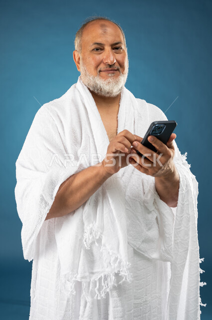 ارتداء ملابس الاحرام، استخدام خدمة تطبيق الارشادات لاداء مناسك الحج والعمرة، بورتريه لرجل مسن مسلم خليجي عربي سعودي يستخدم الهاتف المحمول، تطبيق مرشد الحج الالكتروني، الركن الخامس من أركان الاسلام، موسم الحج 2023، خلفية زرقاء