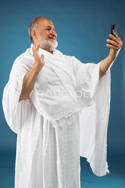 اجراء مكالمة فيديو باستخدام الجوال، بورتريه لرجل مسن مسلم خليجي عربي سعودي يرتدي ملابس الاحرام، بث فيديو مباشر، القاء التحية من خلال شاشة الهاتف المحمول، آداء مناسك الحج والعمرة، الركن الخامس من أركان الاسلام، موسم الحج 2023، خلفية زرقاء