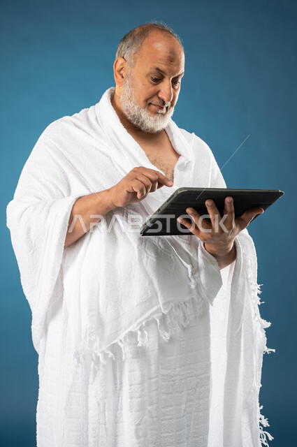استخدام خدمة تطبيق مرشد الحج الالكتروني، بورتريه لرجل مسن مسلم خليجي عربي سعودي يرتدي ملابس الاحرام ويستخدم التابليت المحمول، تطبيق الارشادات لاداء مناسك الحج والعمرة، الركن الخامس من أركان الاسلام، موسم الحج 2023، خلفية زرقاء