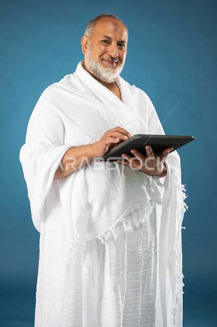 ارتداء ملابس الاحرام، استخدام خدمة تطبيق الارشادات لاداء مناسك الحج والعمرة، بورتريه لرجل مسن مسلم خليجي عربي سعودي يستخدم التابليت المحمول، تطبيق مرشد الحج الالكتروني، الركن الخامس من أركان الاسلام، موسم الحج 2023، خلفية زرقاء
