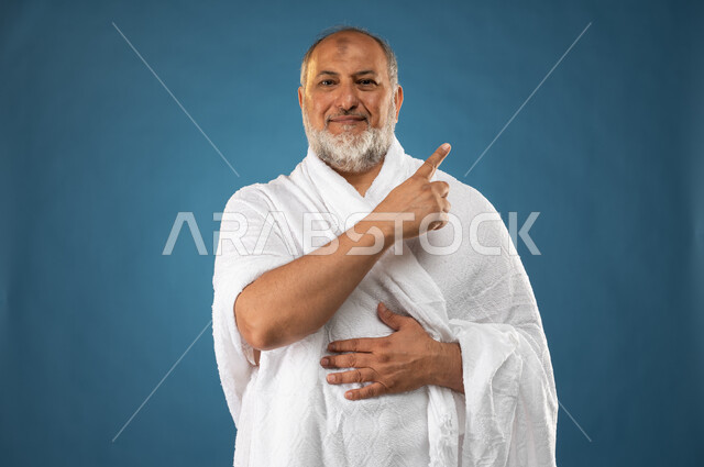 الاشارة باصبع السبابة نحو شيء ما، بورتريه لرجل مسن مسلم عربي سعودي خليجي يرتدي ملابس الاحرام، آداء مناسك الحج والعمرة، ايماءات وجه تدل على السعادة، حركة اصبع واشارات، الركن الخامس من أركان الاسلام، موسم الحج 2023، خلفية زرقاء