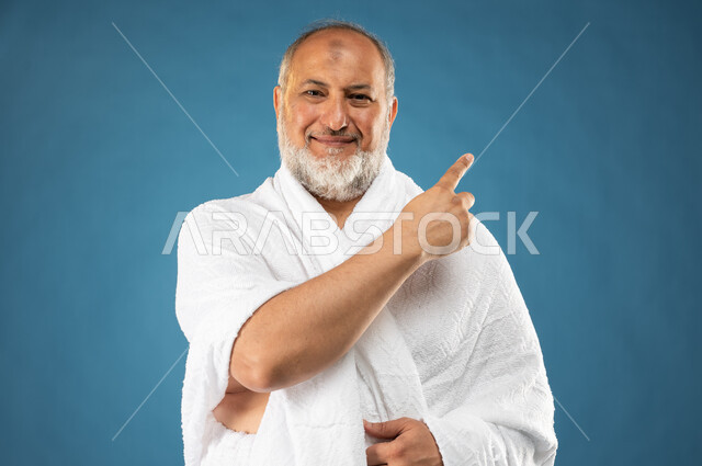 حركة اصبع واشارات، آداء مناسك الحج والعمرة، بورتريه لرجل مسن مسلم عربي سعودي خليجي يرتدي ملابس الاحرام ويشير باصبع السبابة نحو شيء ما، ايماءات وجه تدل على السعادة، الركن الخامس من أركان الاسلام، موسم الحج 2023، خلفية زرقاء