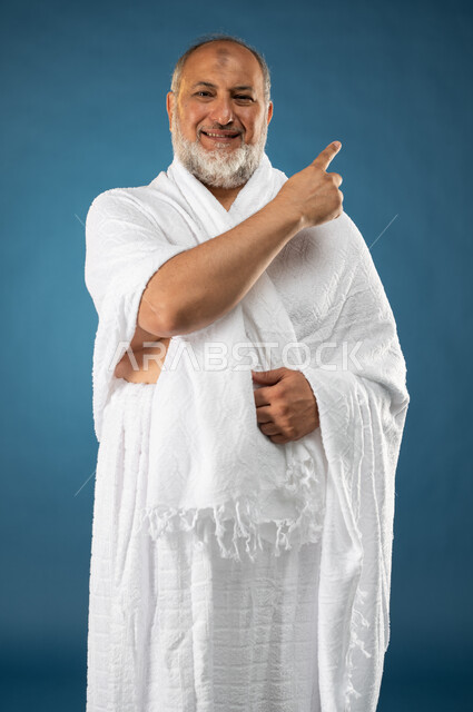 Performing the rituals of Hajj and Umrah, a portrait of an elderly Muslim, Arab, Saudi, Gulf man wearing ihram clothes and pointing with his index finger towards something, facial gestures indicating happiness, finger movement and gestures, the fifth pill