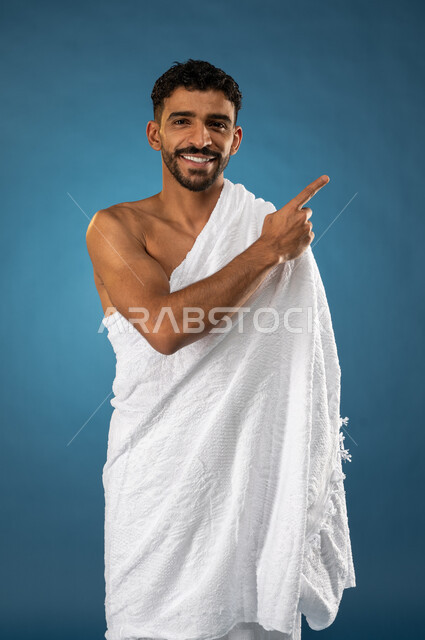 حركة اصبع واشارات، آداء مناسك الحج والعمرة، بورتريه لشاب مسلم عربي سعودي خليجي يرتدي ملابس الاحرام ويشير باصبع السبابة نحو شيء ما، ايماءات وجه تدل على السعادة، الركن الخامس من أركان الاسلام، موسم الحج 2023، خلفية زرقاء