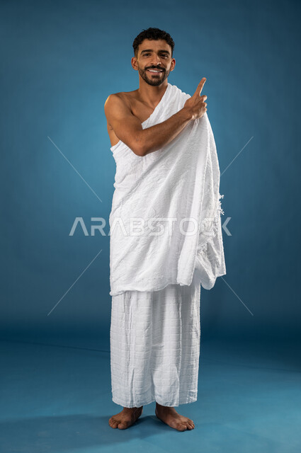 الاشارة باصبع السبابة نحو شيء ما، بورتريه لشاب مسلم سعودي خليجي يرتدي ملابس الاحرام، آداء مناسك الحج والعمرة، ايماءات وجه تدل على السعادة، حركة اصبع واشارات، الركن الخامس من أركان الاسلام، موسم الحج 2023، صورة الجسم بالطول الكامل، خلفية زرقاء