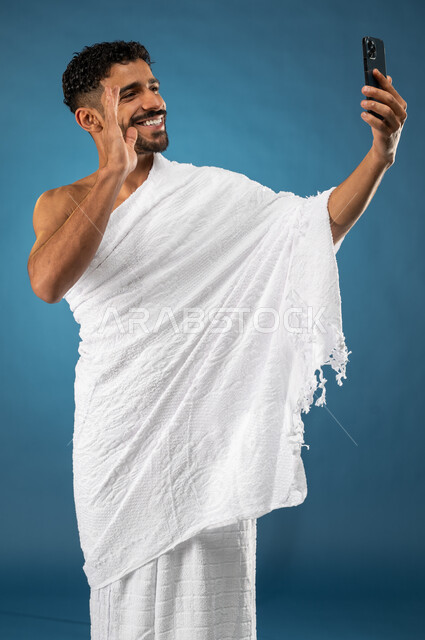 اجراء مكالمة فيديو باستخدام الجوال، بورتريه لشاب مسلم خليجي عربي سعودي يرتدي ملابس الاحرام، بث فيديو مباشر، القاء التحية من خلال شاشة الهاتف المحمول، آداء مناسك الحج والعمرة، الركن الخامس من أركان الاسلام، موسم الحج 2023، خلفية زرقاء