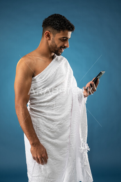 استخدام خدمة تطبيق الدليل لأداء مناسك الحج والعمرة ، صورة لشاب سعودي خليجي مسلم يرتدي ملابس الإحرام ويستخدم الهاتف المحمول ، تطبيق دليل الحج الإلكتروني ، الركن الخامس من الإسلام ، موسم الحج 2023