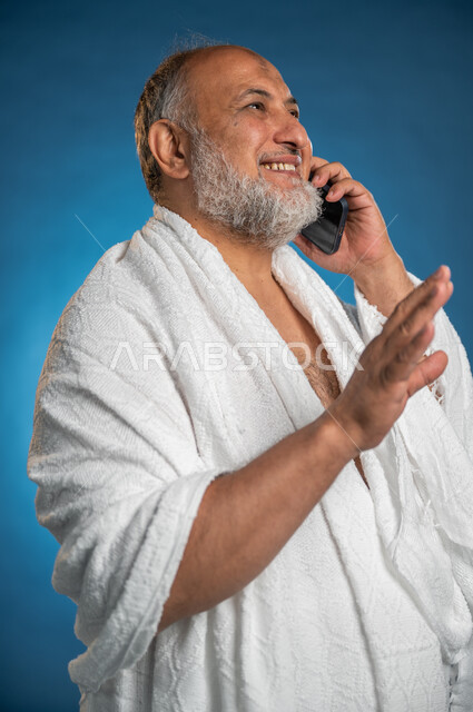 استخدام الهاتف المحمول، بورتريه لرجل مسن مسلم خليجي عربي سعودي يرتدي ملابس الاحرام ويجري مكالمة هاتفية، التواصل الاجتماعي والعلاقات الاجتماعية، آداء مناسك الحج والعمرة، الركن الخامس من أركان الاسلام، موسم الحج 2023، خلفية زرقاء