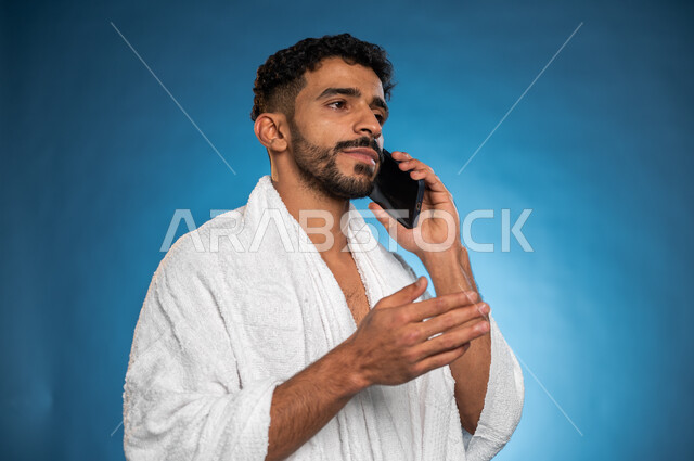 Social communication and social relations, a portrait of a Saudi Arab Gulf Muslim man wearing ihram clothes and making a phone call, using a mobile phone, performing Hajj and Umrah rituals, the fifth pillar of Islam, Hajj season 2023, blue background