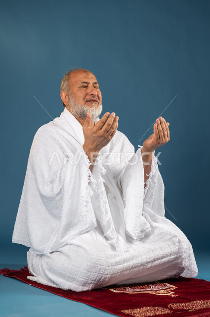آداء مناسك الحج والعمرة، بورتريه لرجل مسن مسلم عربي سعودي خليجي يرتدي ملابس الاحرام ويرفع يديه لاعلى، الجلوس على سجادة الصلاة والدعاء والعباده والتقرب الى الله، الركن الخامس من أركان الاسلام، موسم الحج 2023، خلفية زرقاء